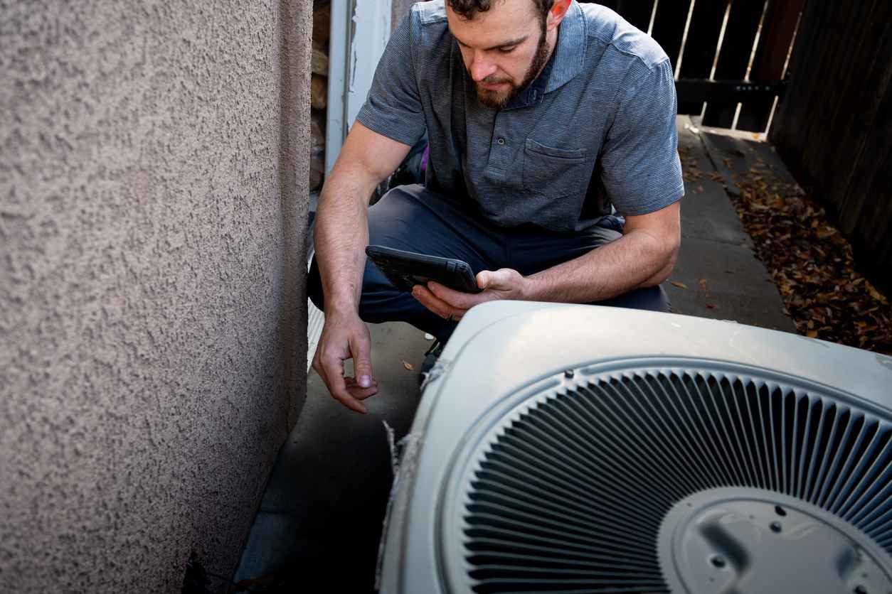 Young Male Property Inspector Photographing a Line to a Residential Air Conditioner Condenser Unit at the side of a stucco home