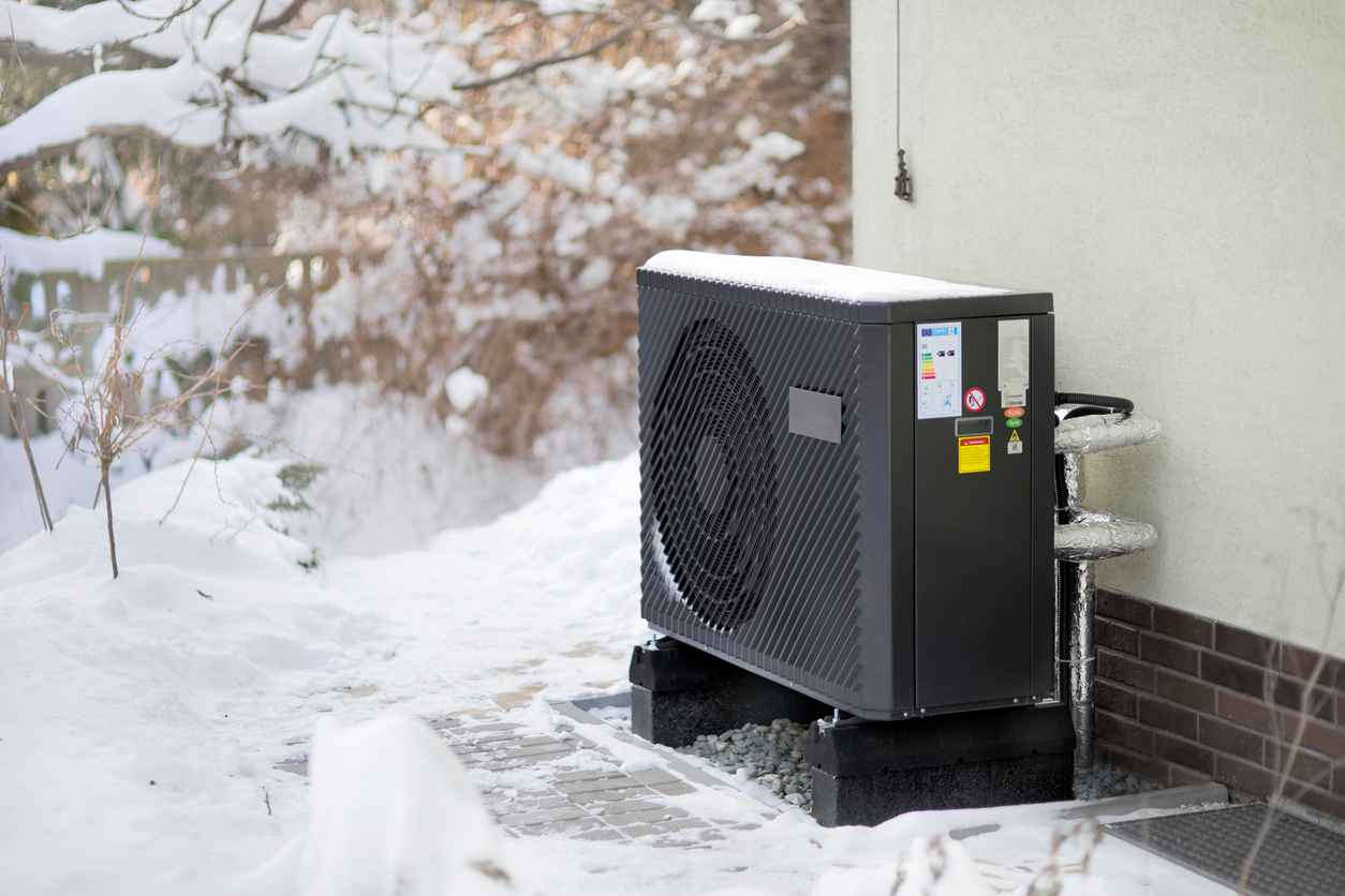 Photo of a modern air source heat pump unit standing against the residential building wall. Shot in winter, in a snowy setting.