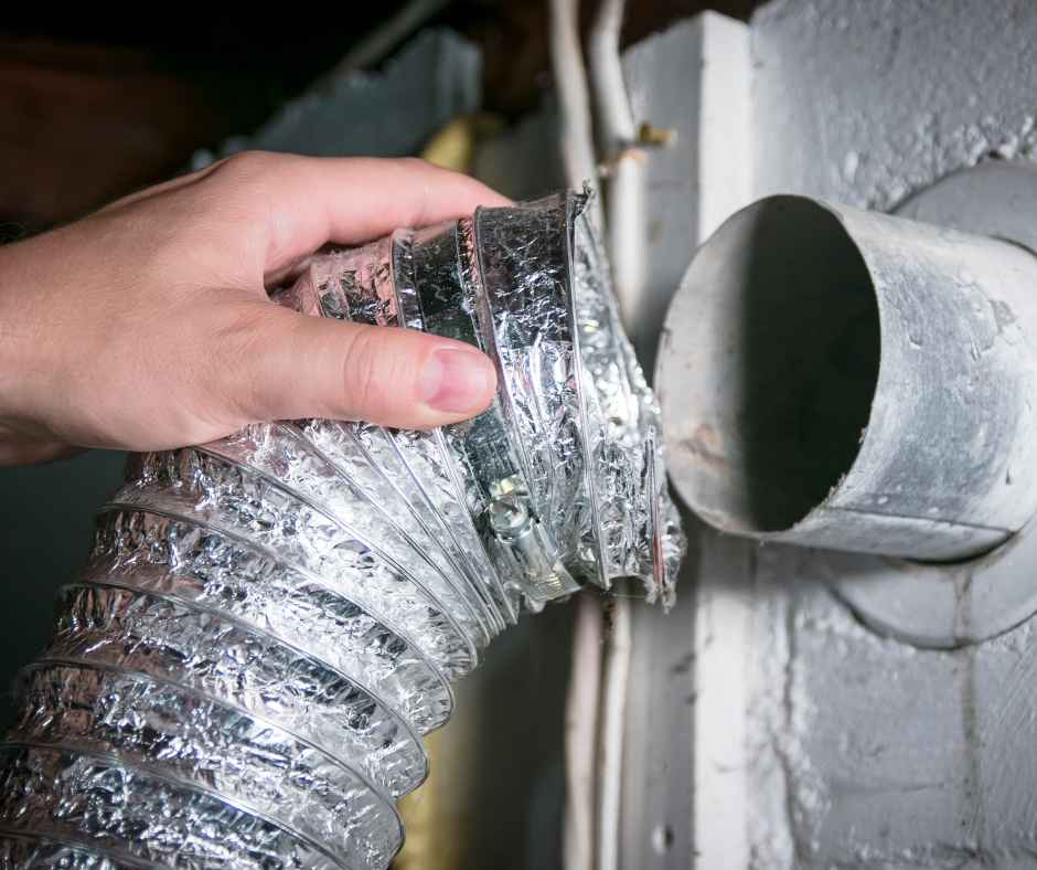 Hand holding a silver flexible aluminum HVAC duct being inserted into a round metal vent opening in a gray wall.